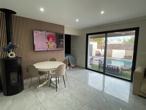 Ein modernes Wohnzimmer mit flachbildfernseher an der Wand, einem Essbereich mit rundem Tisch und Stühlen, einem schwarzen Kaminofen und einer Glastür, die zu einem Garten mit Pool führt.