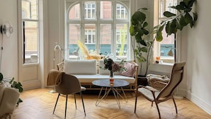 Een kamer met een visgraatvloer, een witte bank, een ronde tafel en een grote plant.