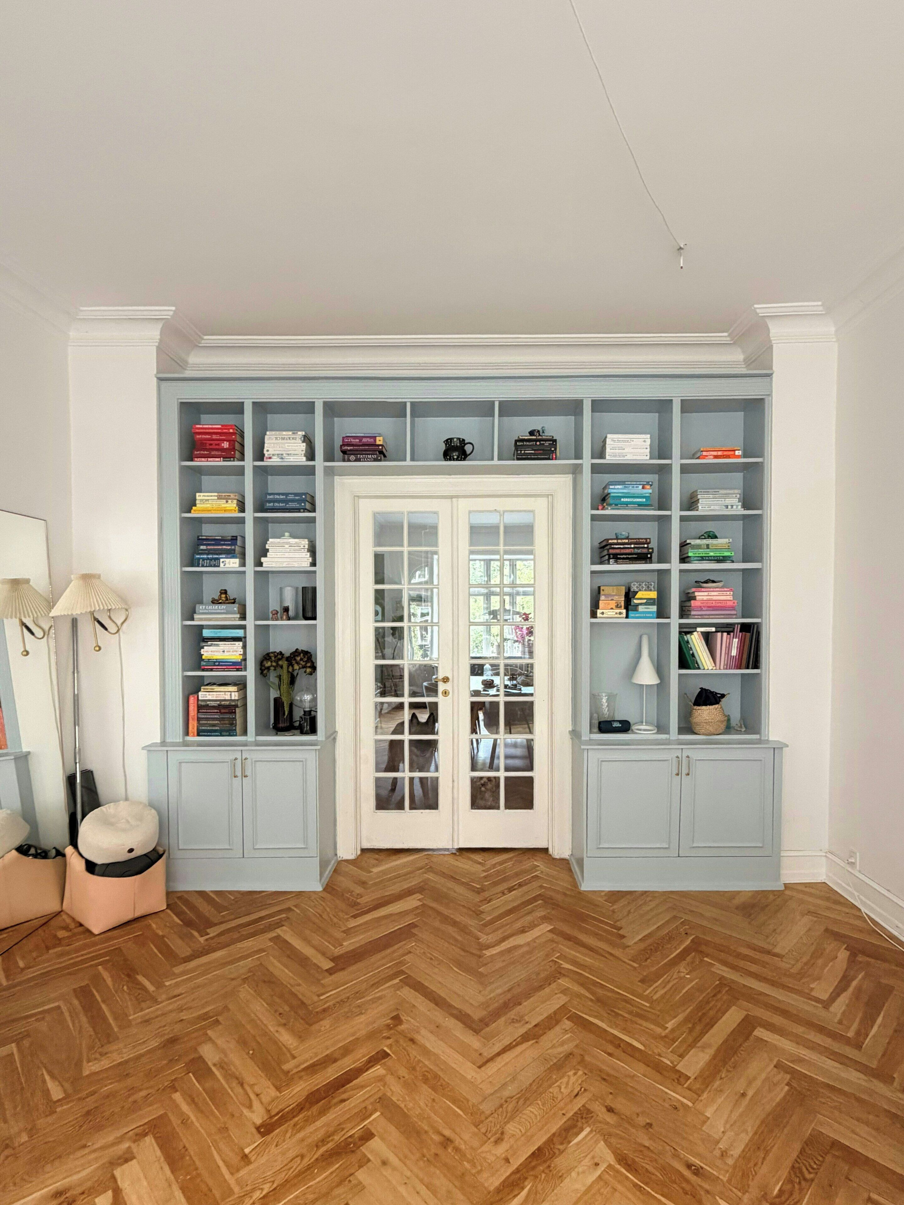 A room with a herringbone wood floor, a white door with glass panels, and a light blue built-in bookshelf with various items.