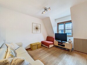 A modern living room with a sofa, a red chair, a television, and a window with blinds. - "Emmy" Apartment – Oostduinkerke – Cozy vacation apartment near the beach (Koksijde)