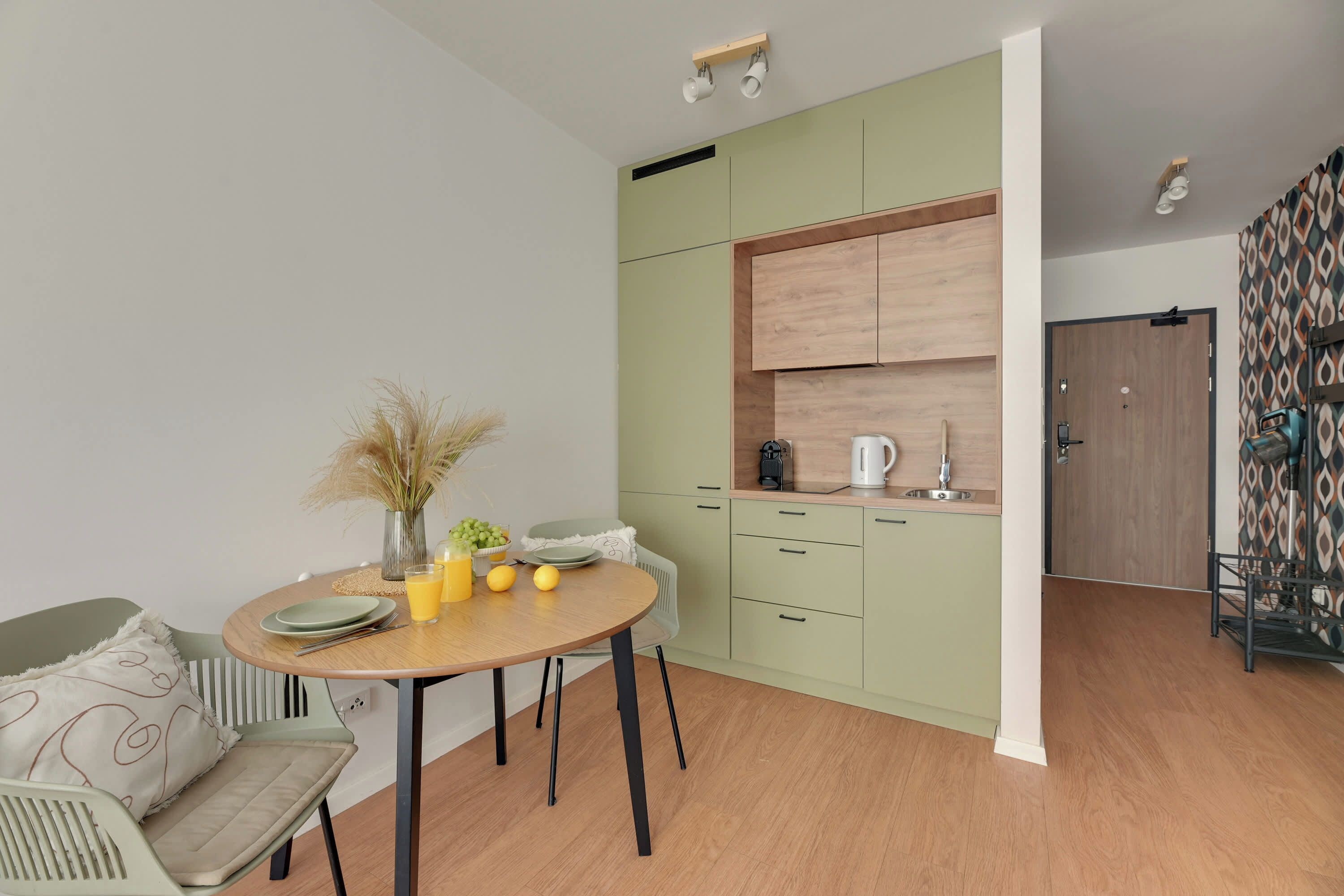 A compact kitchen with a dining area, featuring a round table, chairs, and a small cabinet with a kettle.