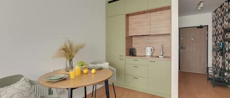 A compact kitchen with a dining area, featuring a round table, chairs, and a small cabinet with a kettle.