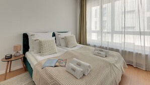 Una cama bien hecha con una colcha texturizada, una mesita de noche con una lámpara y una ventana con cortinas traslúcidas.