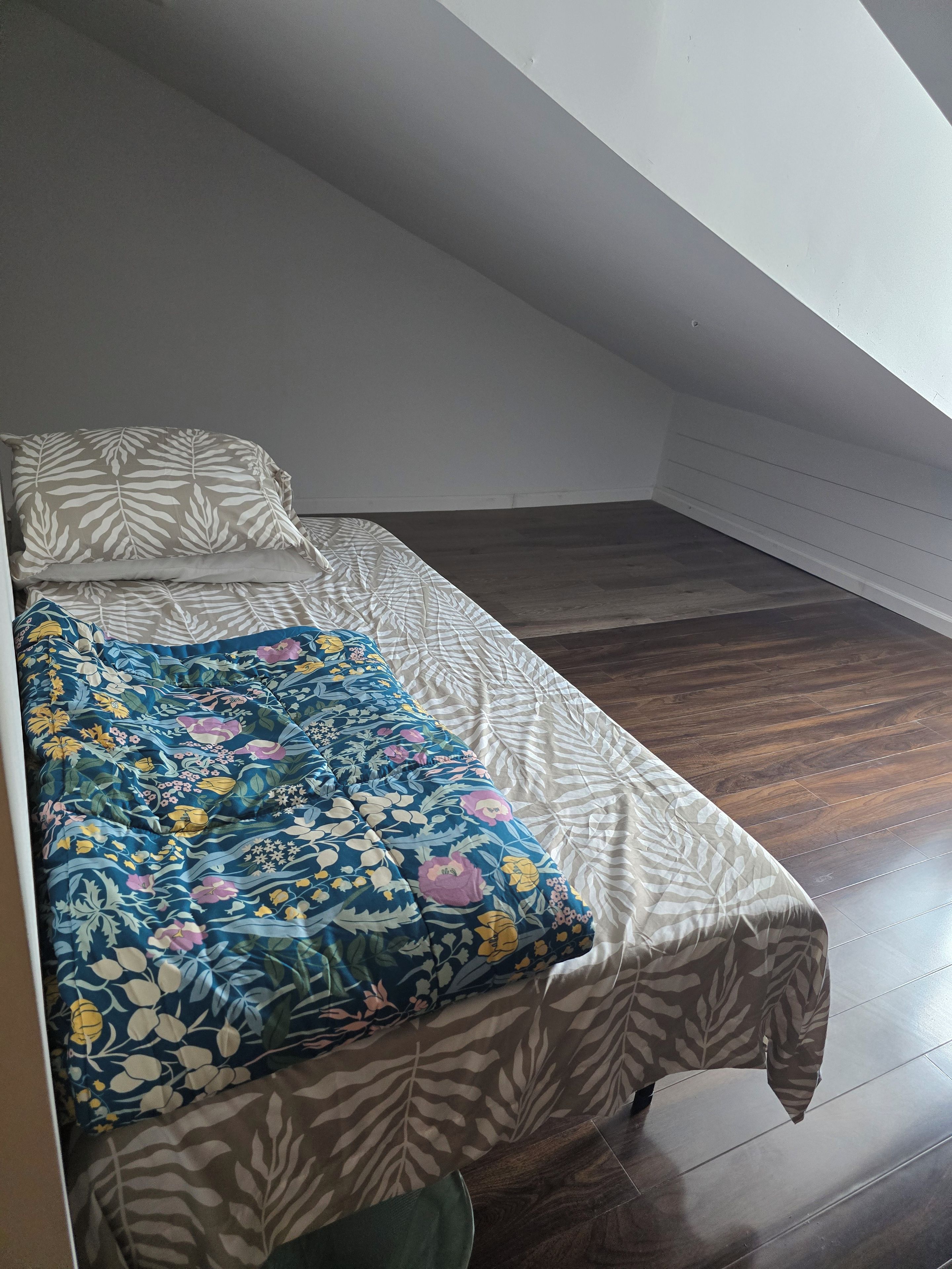 Una habitación pequeña con cama individual, cubrecama estampado y almohada lisa. El piso es de madera y el techo, inclinado.