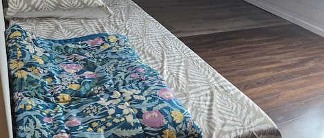 Una camera piccola con un letto singolo, una coperta a motivi e un cuscino semplice. La stanza ha pavimento in legno e un soffitto spiovente.