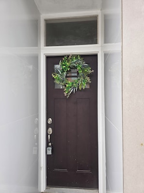 Une porte brun foncé ornée d’une guirlande, avec une fenêtre au-dessus et un carillon.