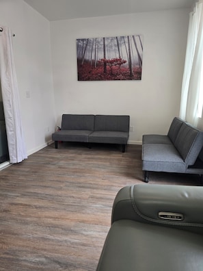 Un salon avec un canapé gris, un canapé vert et un tableau représentant une forêt.