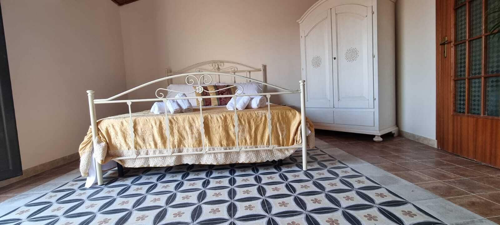Uma cama com estrutura de metal e colcha estampada, um guarda-roupa branco e um chão com padrão.