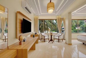 Habitación de hotel moderna con televisión de pantalla plana, escritorio de madera, dos sillas, una mesa redonda y ventanas amplias con cortinas.