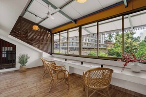 Una habitación con un gran ventanal, sillas de mimbre, un jarrón blanco con flores rojas y un ventilador de techo.