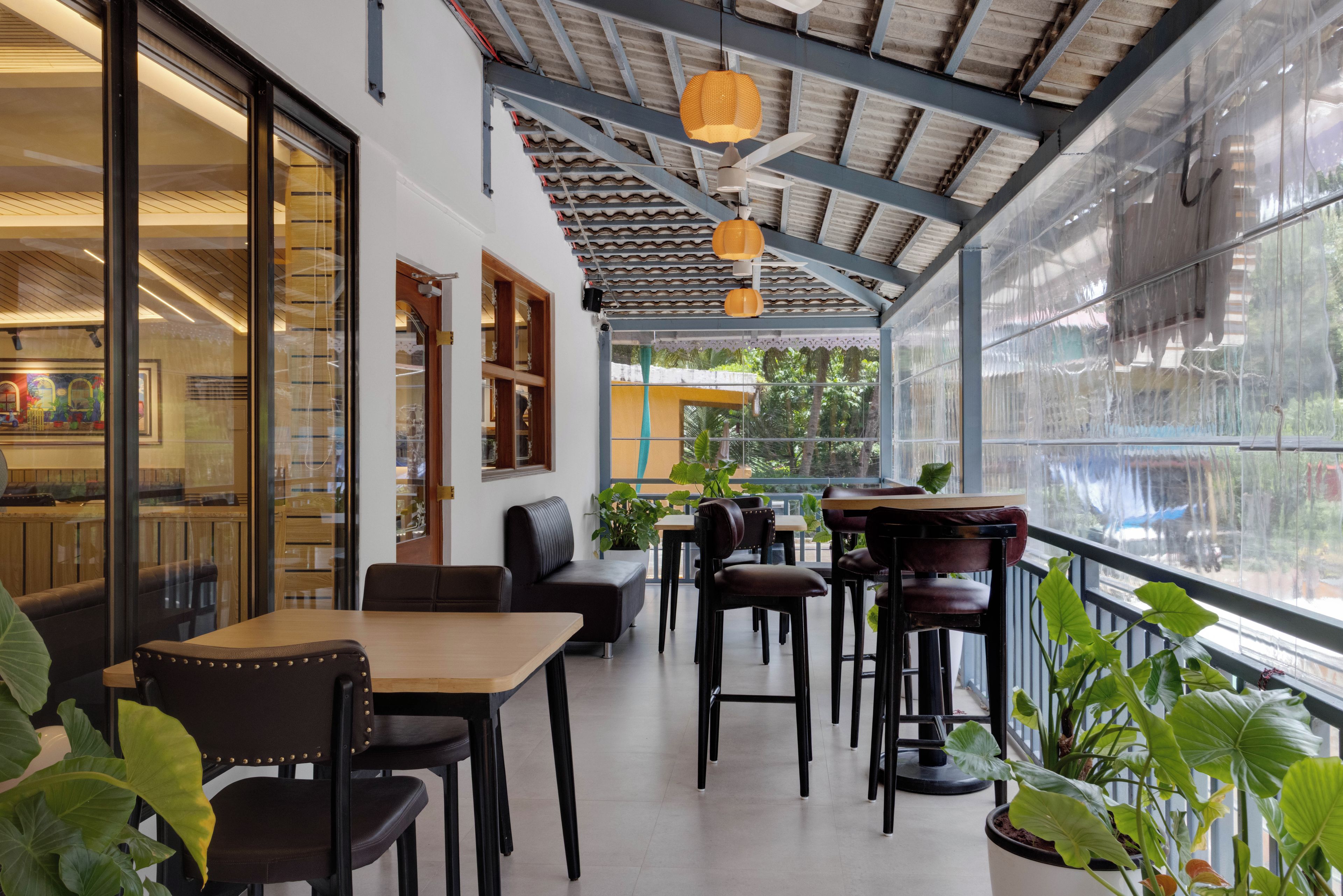 A covered outdoor seating area with tables and chairs, large windows, and potted plants.