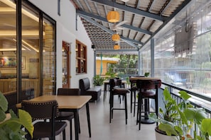 Un espacio al aire libre con techo, mesas y sillas, ventanas amplias y plantas en macetas.