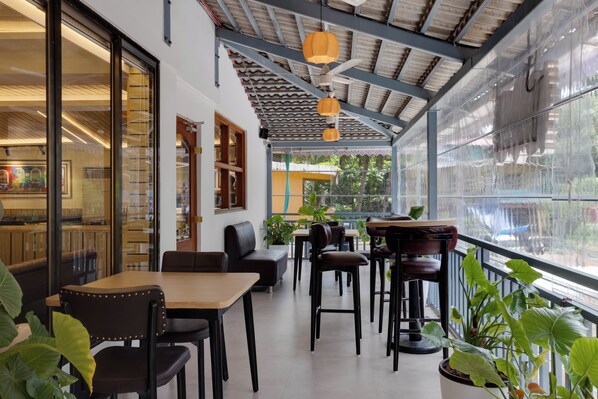 Un espacio al aire libre con techo, mesas y sillas, ventanas amplias y plantas en macetas.