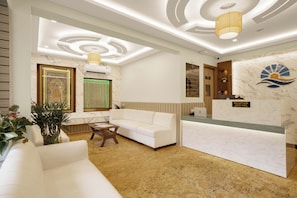 A hotel reception area with a white reception desk, a white sofa, a wooden coffee table, and a large framed picture on the wall.