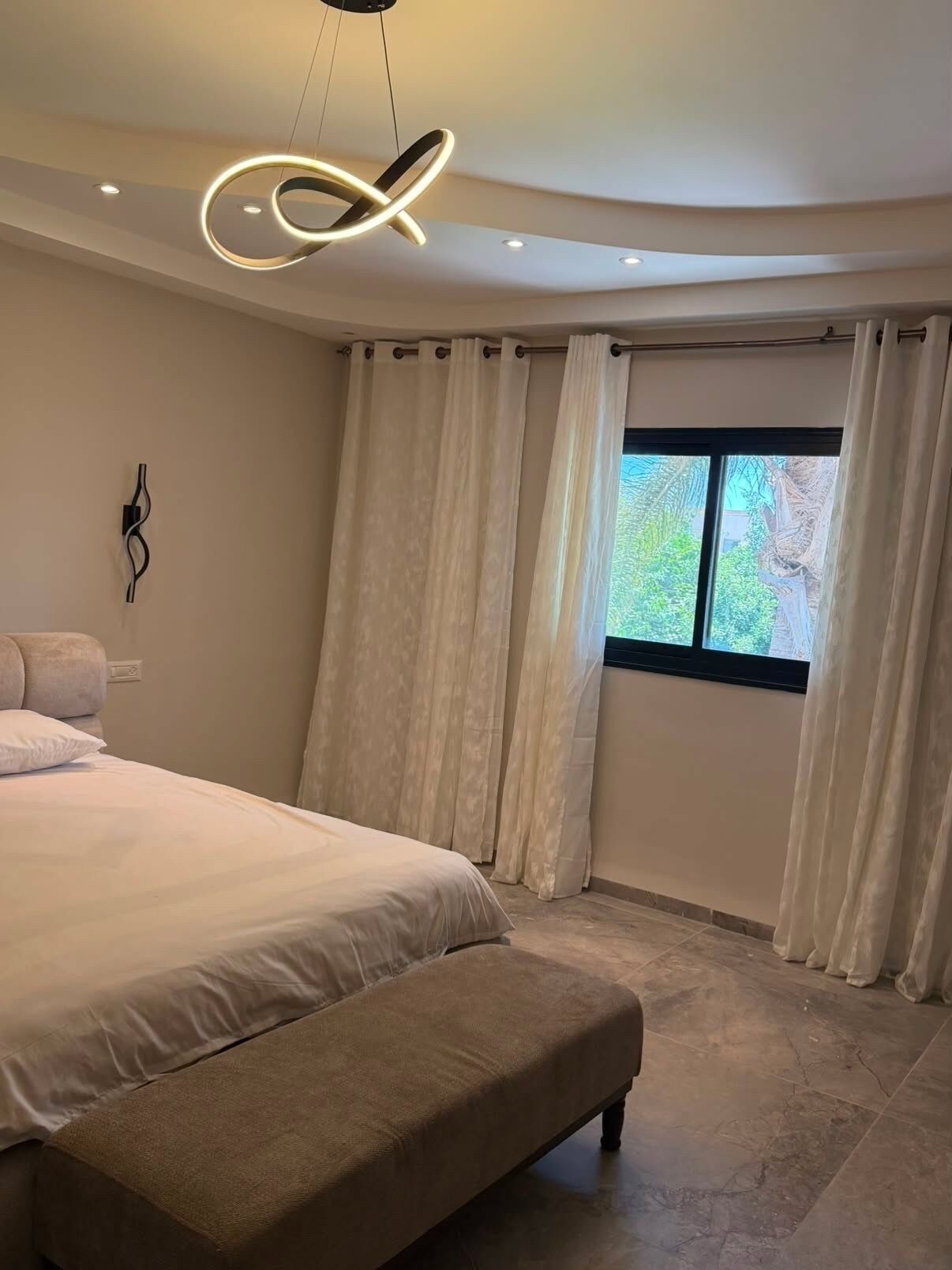 A bedroom with a large bed, a bench, a modern ceiling light, and a window with curtains.