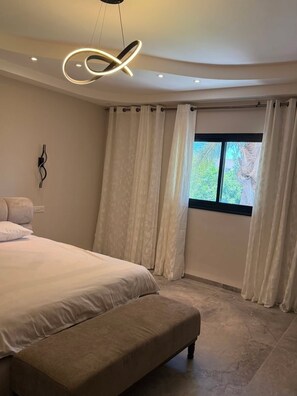 A bedroom with a large bed, a bench, a modern ceiling light, and a window with curtains.
