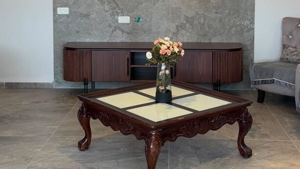 A room with a large, ornate wooden coffee table, a modern chandelier, and a grey stone wall.