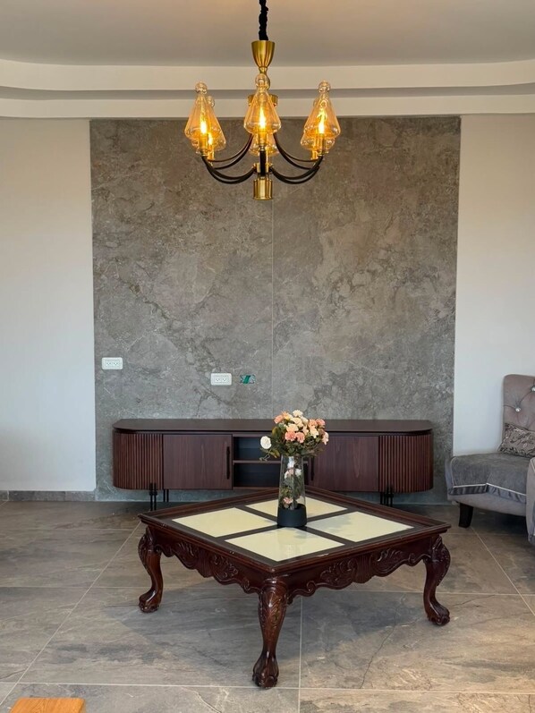 A room with a large, ornate wooden coffee table, a modern chandelier, and a grey stone wall.