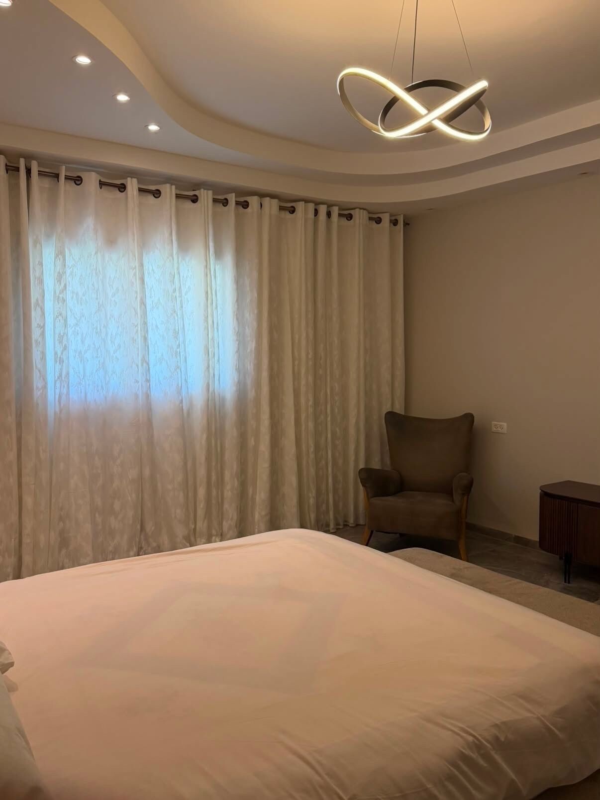 Quarto de hotel com uma cama grande, uma cadeira, uma escrivaninha e um lustre moderno.