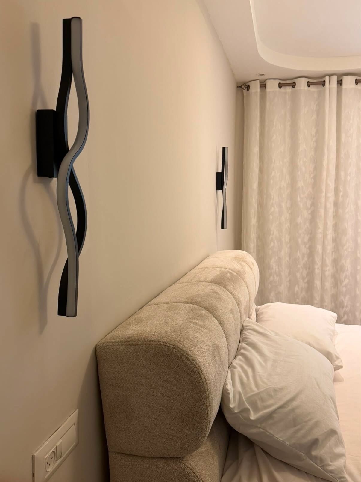 A hotel room with a bed, a wall-mounted light fixture, and a power outlet.