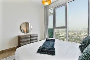A modern bedroom with a large bed, a dresser, a round mirror, and a view of a cityscape through large windows. - Elegant 3 BR | Wasl 1 (Dubai)