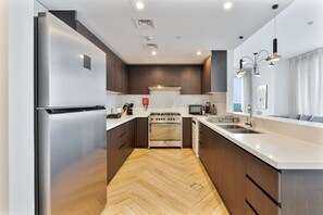 A modern kitchen with stainless steel appliances, a herringbone floor pattern, and a large island with a sink. - Elegant 3 BR | Wasl 1 (Dubai)