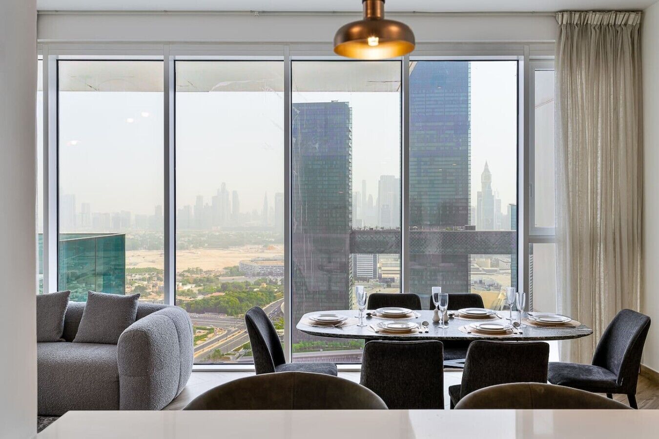 Un coin repas avec une table dressée pour quatre personnes, de grandes fenêtres offrant une vue sur la ville et un luminaire suspendu moderne.