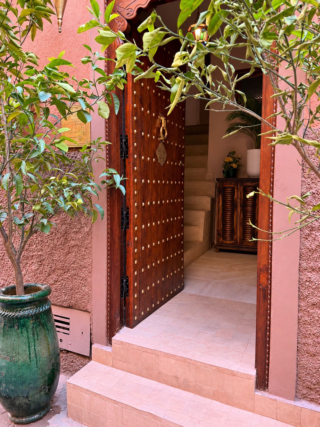 Une porte en bois ornée de motifs décoratifs et dotée d’une poignée en laiton, qui donne sur un escalier et une pièce contenant un meuble et une plante en pot.