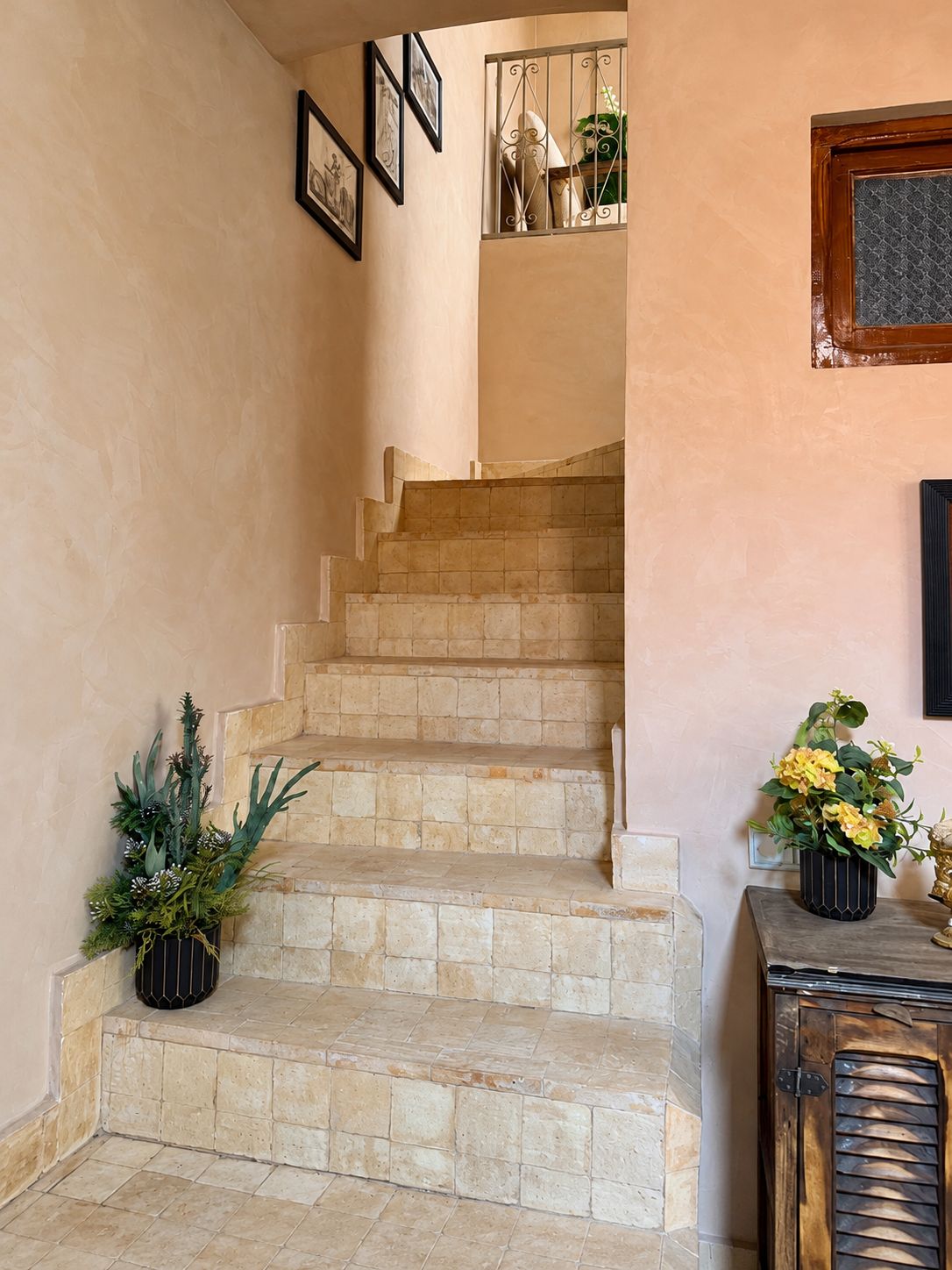 A staircase with stone steps leading to an upper floor, flanked by potted plants and framed artwork.