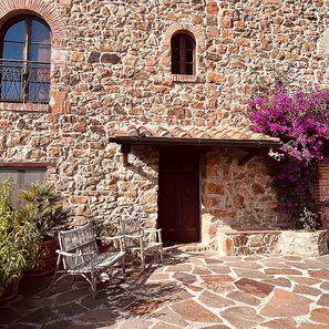 Ein Steinhaus mit Ziegeldach, einer Holztür und einem kleinen Fenster. Vor der Tür stehen zwei Korbstühle und ein Blumentopf.