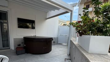 A modern outdoor patio with a hot tub, a seating area, and a white planter with plants.