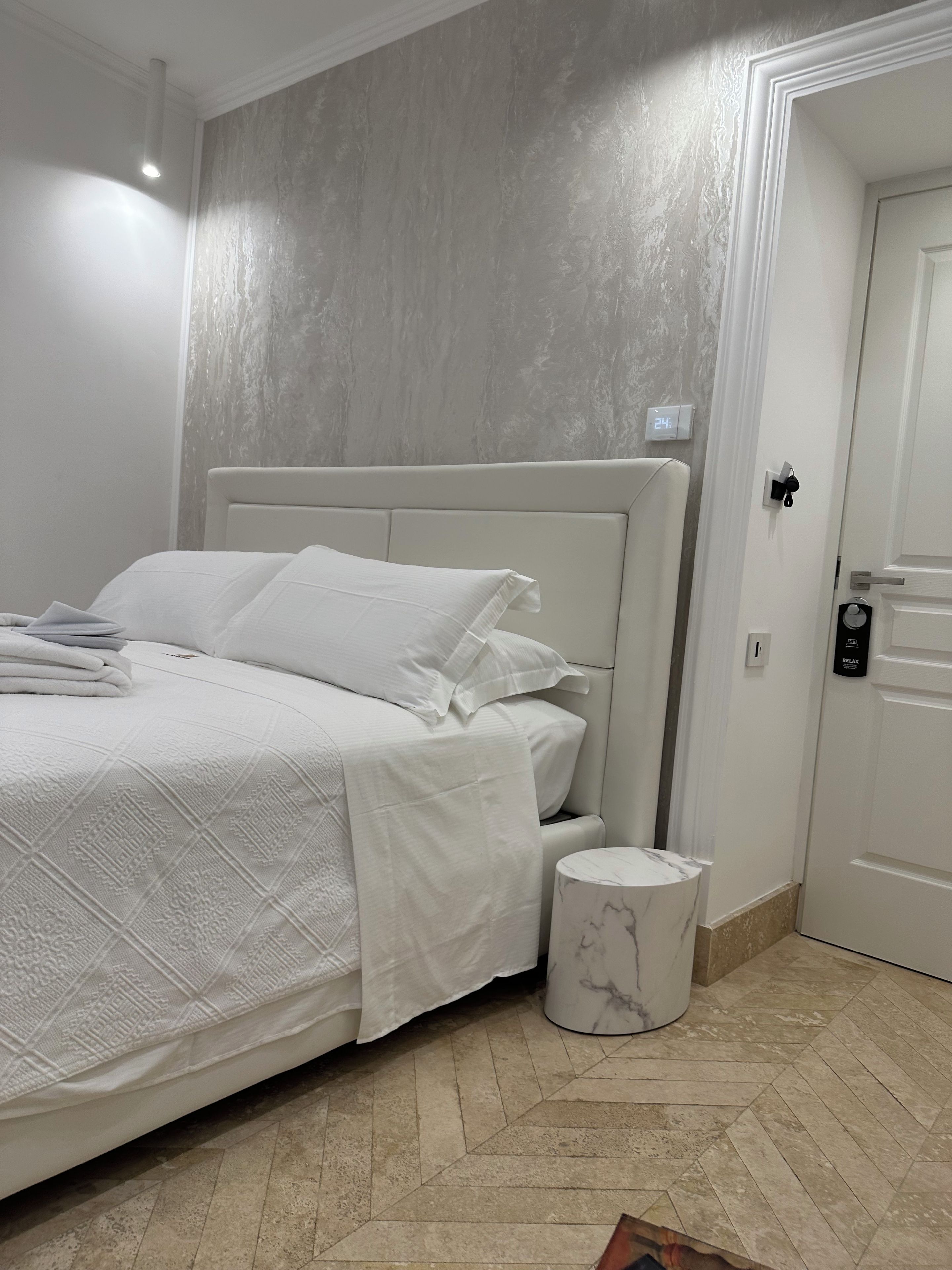 Una cama bien tendida con cabecera blanca, una mesita de noche de mármol y una puerta blanca con cerradura.