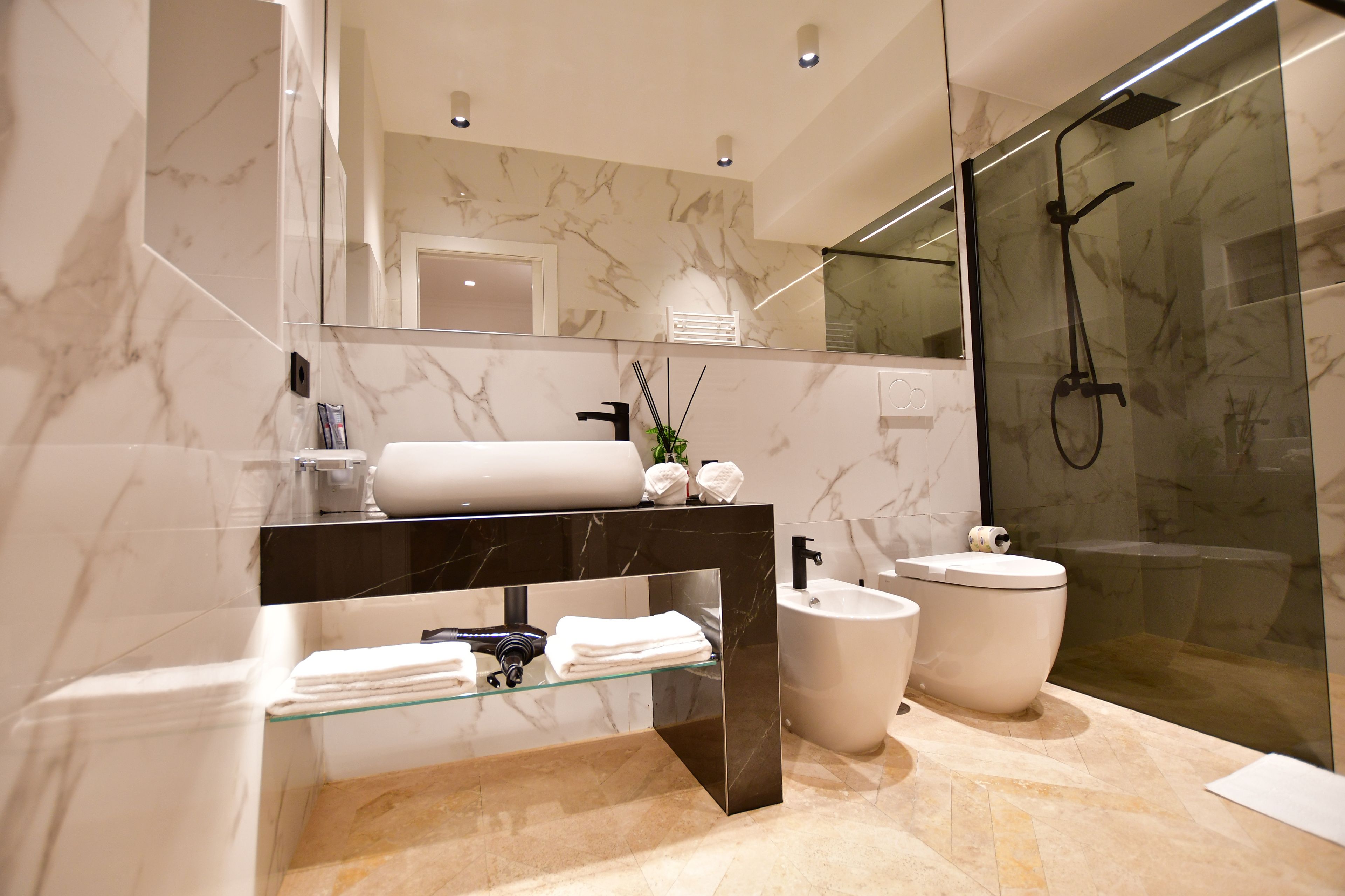 Un bagno moderno con box doccia in vetro, parete rivestita in marmo, ripiano in vetro con articoli da toilette e mobile lavabo nero.