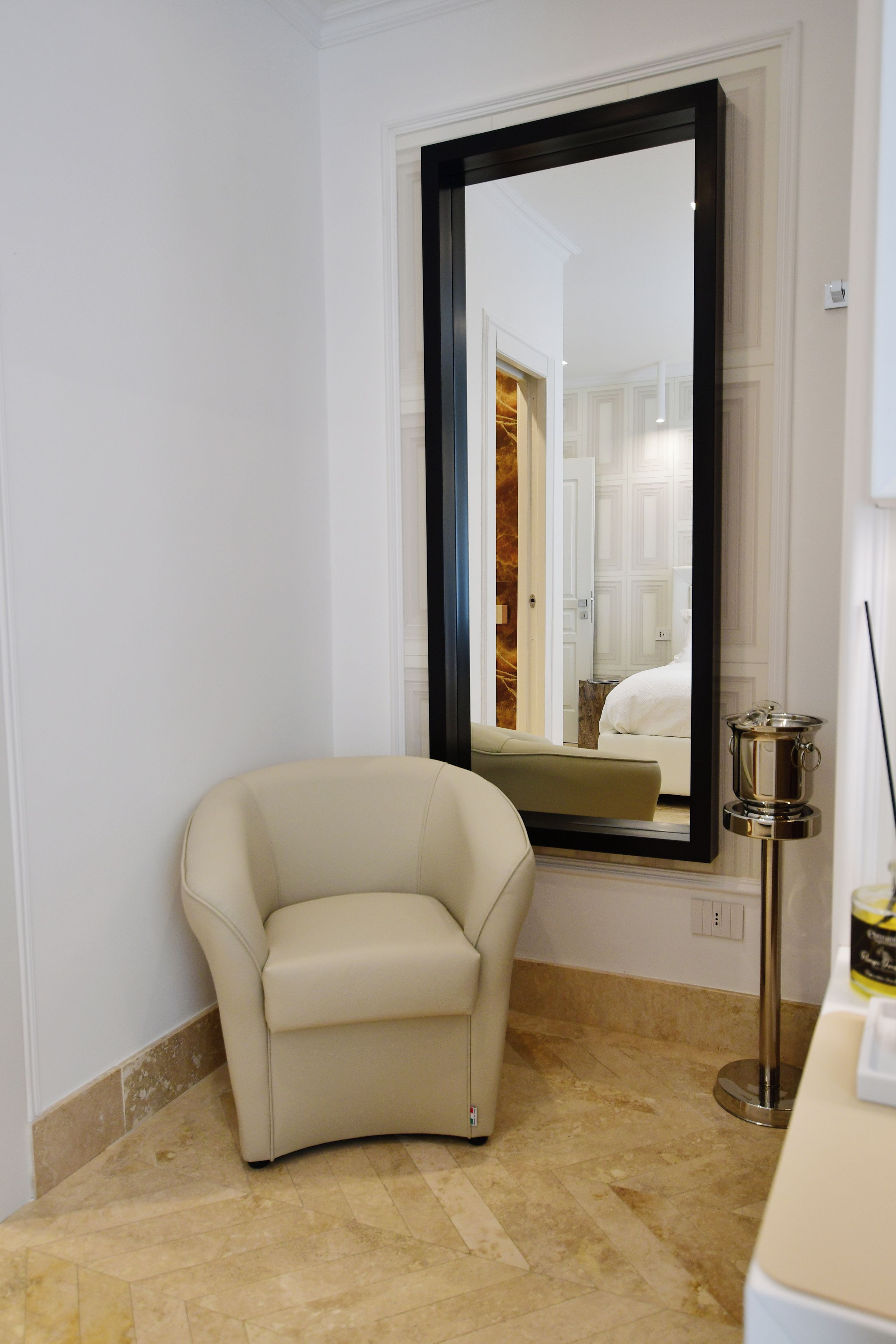 Una habitación con un espejo grande, un sillón color beige, una lámpara de pie y una mesita con una botella.