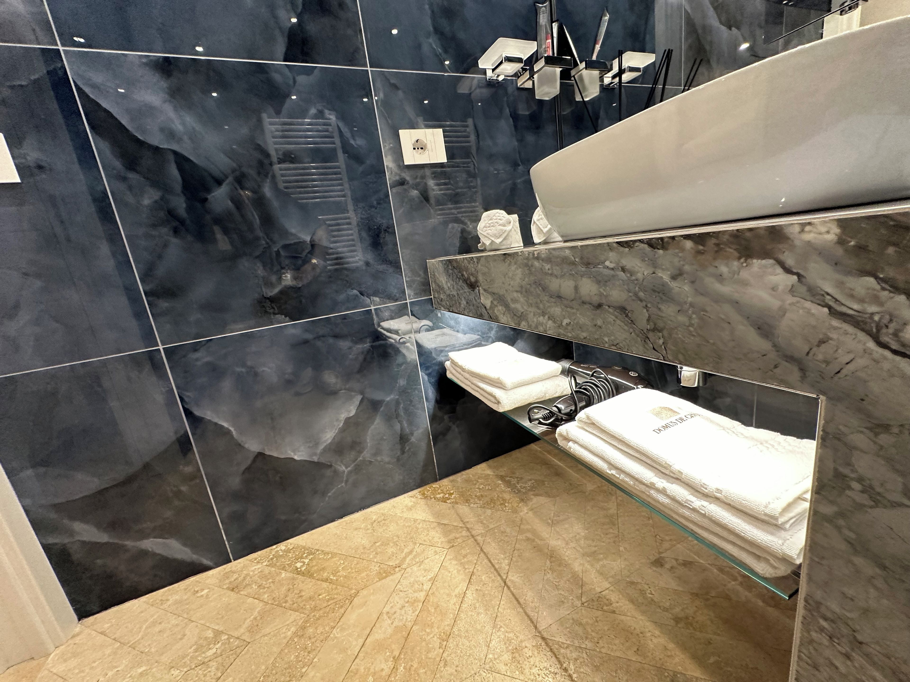 Une salle de bain moderne avec un meuble-lavabo en marbre, une étagère en verre où sont posées des serviettes et un miroir.