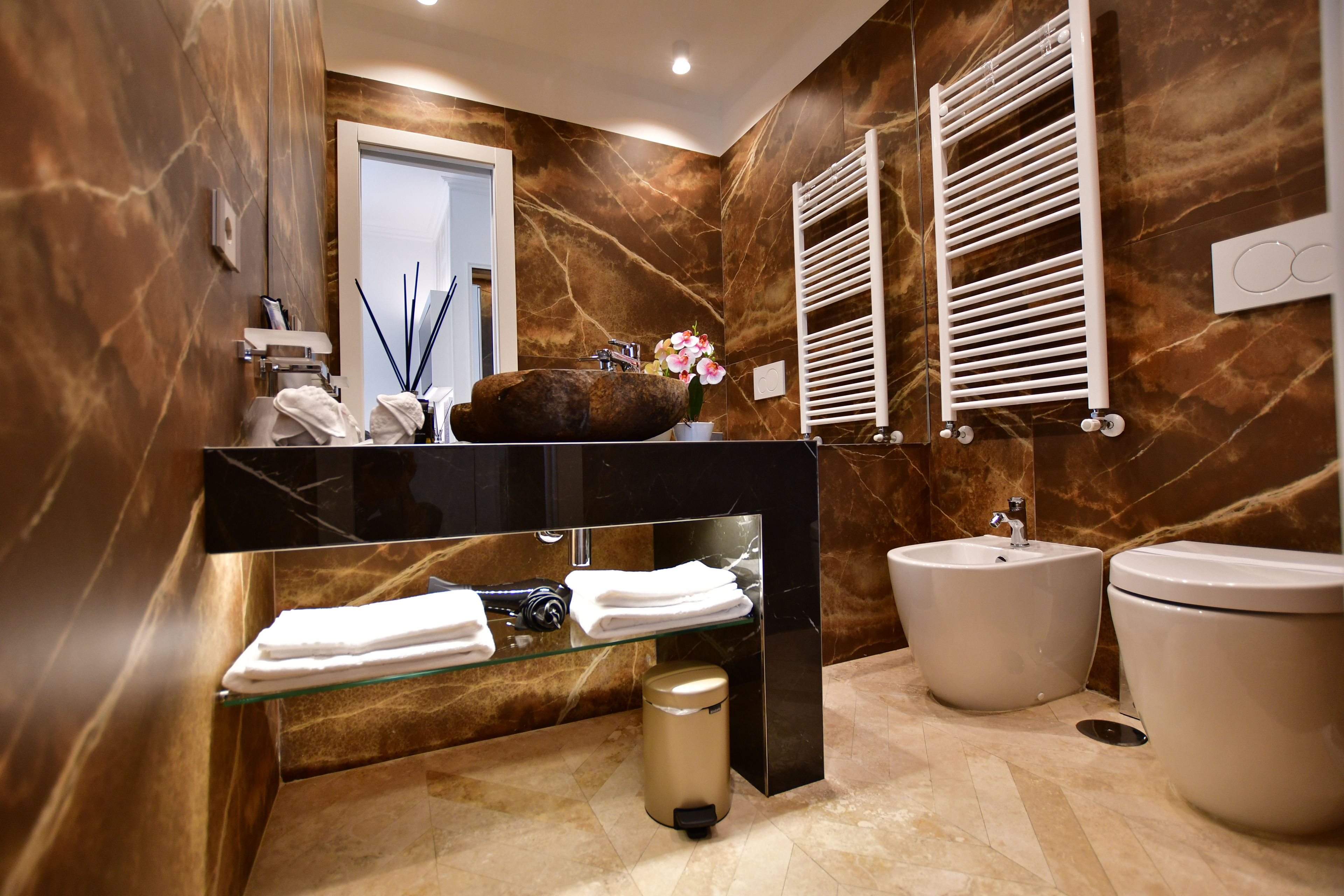 Une salle de bain moderne avec un mur en marbre, une étagère en verre où sont posées des serviettes, un lavabo et des toilettes.