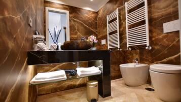 Une salle de bain moderne avec un mur en marbre, une étagère en verre où sont posées des serviettes, un lavabo et des toilettes.