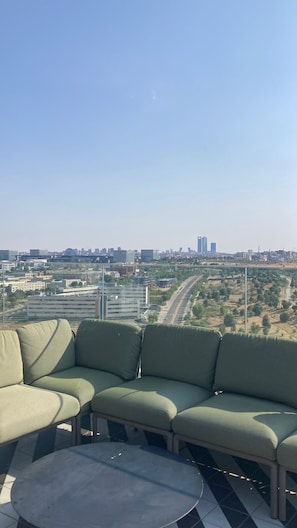 A rooftop terrace with a green sofa and a round table, overlooking a cityscape with buildings and a road. - Apartment for Formula 1 (Alcobendas)