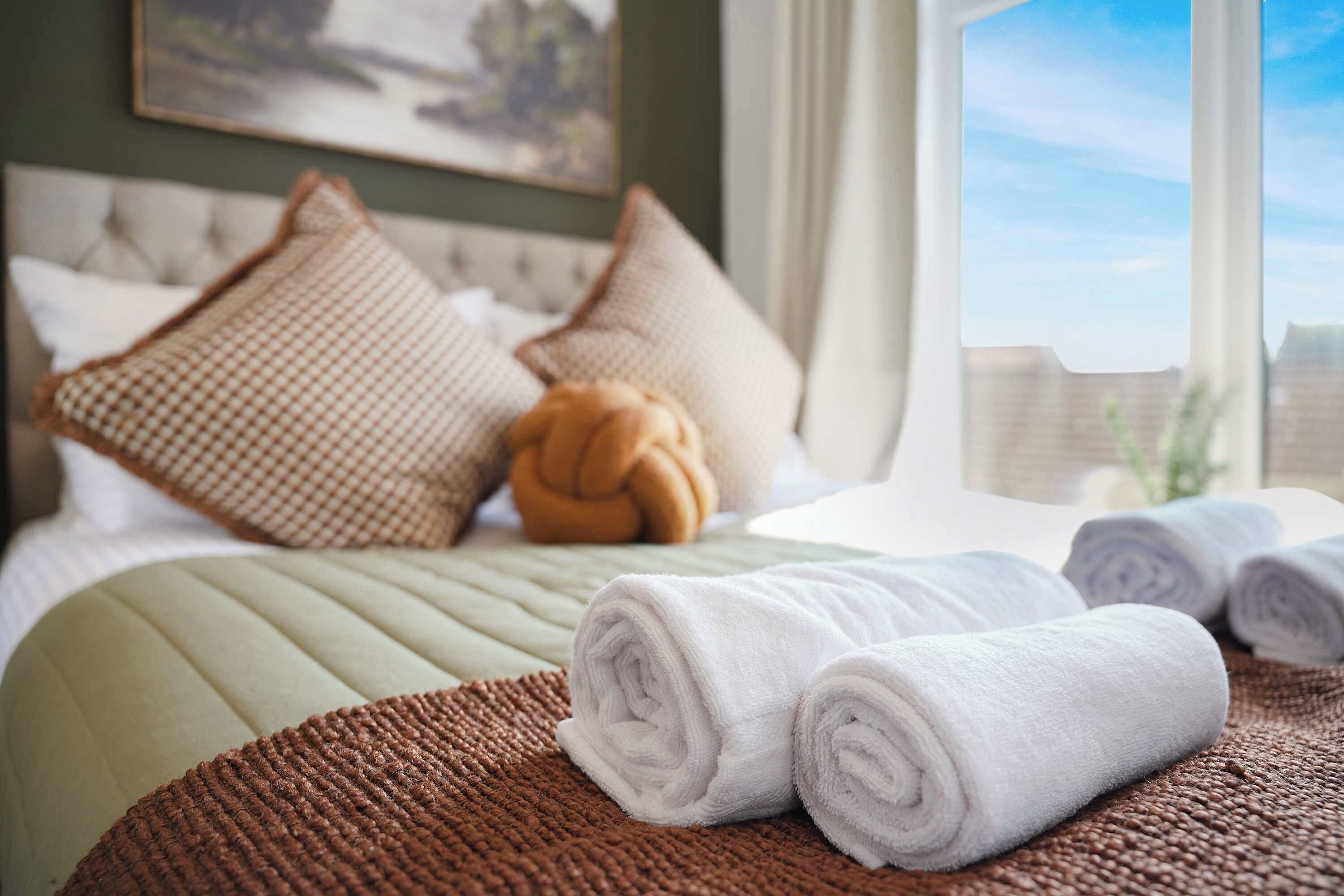 Una cama bien hecha con toallas blancas enrolladas, una manta marrón tejida y almohadones decorativos.