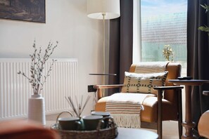 Un salon moderne avec une grande fenêtre, un lampadaire, un radiateur et un fauteuil agrémenté d’un plaid.