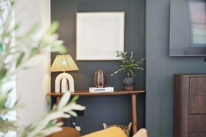 Ein modernes Wohnzimmer mit dunkler Wand, einem gerahmten abstrakten Kunstwerk, einer Lampe, einer Vase und einem kleinen Tisch mit der Aufschrift 'LONDON'.