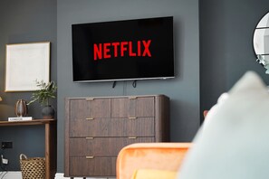 A modern living room with a flat-screen TV mounted on the wall displaying 'NETFLIX' in red letters, a wooden cabinet with drawers, a small plant, and a framed document on a shelf. - Spacious 4BR near Hornsea Museum | Sleeps 8+WiFi (Hornsea)
