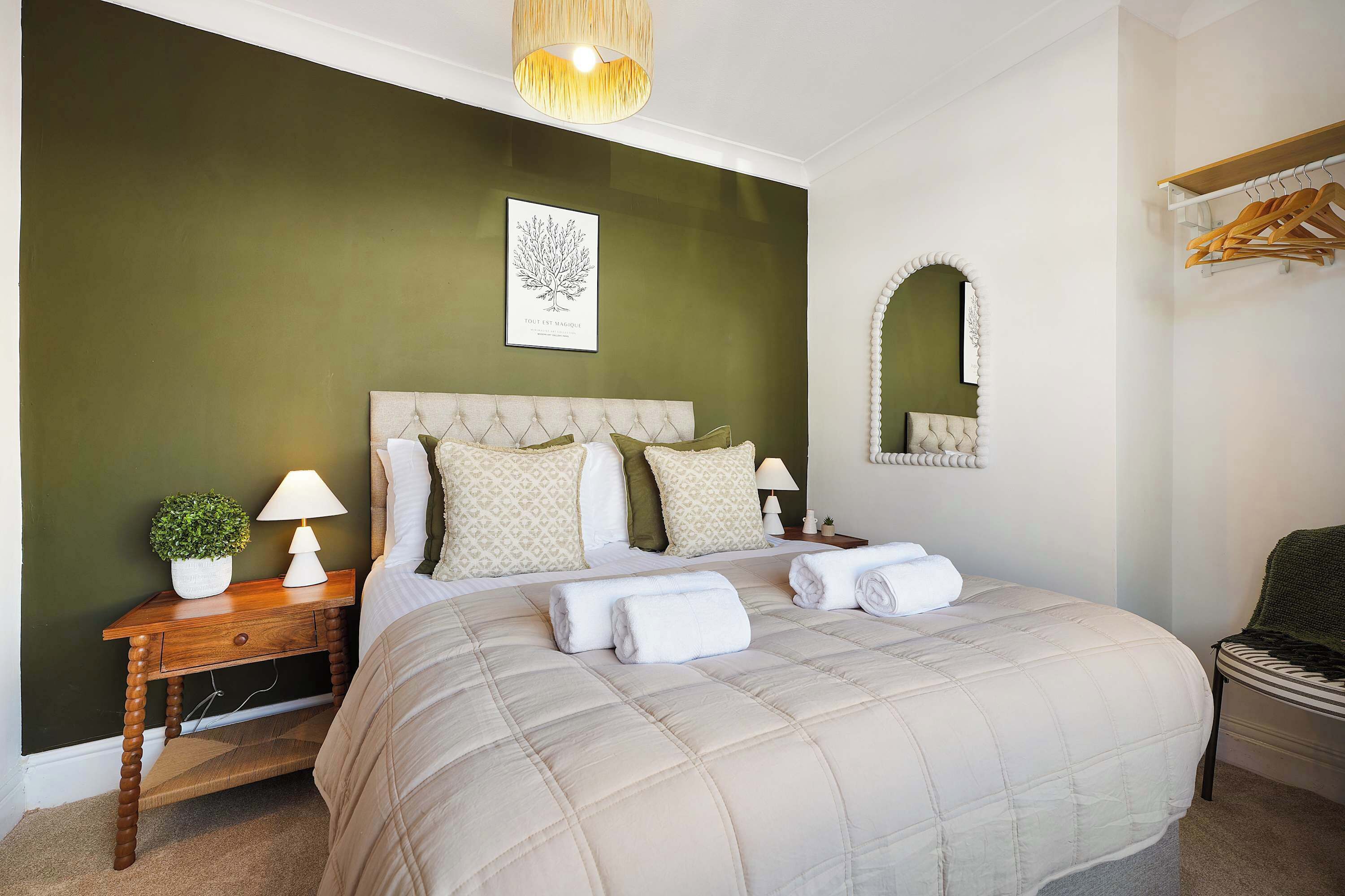 A bedroom with a large bed, a bedside table with a lamp, a small plant, and a mirror.