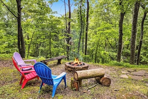 Ein Waldcampingplatz mit Feuerstelle, bunten Stühlen und einem Baumstamm.
