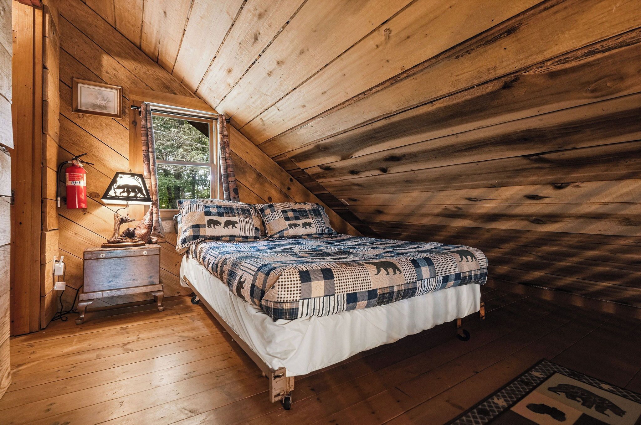 Ein gemütliches Schlafzimmer mit schrägen Wänden, einem Bett mit einem gemusterten Bettbezug, einem Nachttisch mit Lampe und einem Fenster mit Blick auf Bäume.
