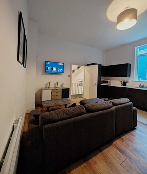 A modern living room with a sectional sofa, a flat-screen TV, a dining table, and a kitchen area with a microwave. - Unique 4BR City Apartment | Terrace + Sleeps 10 (Liverpool)