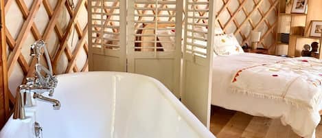 Una habitación con una bañera grande, paredes de madera celosía, una cama con cubrecama blanco y un estante con diversos objetos.