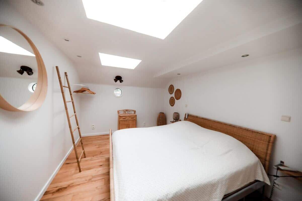 Een slaapkamer met dakraam, houten ladder, bed, dressoir en spiegel.