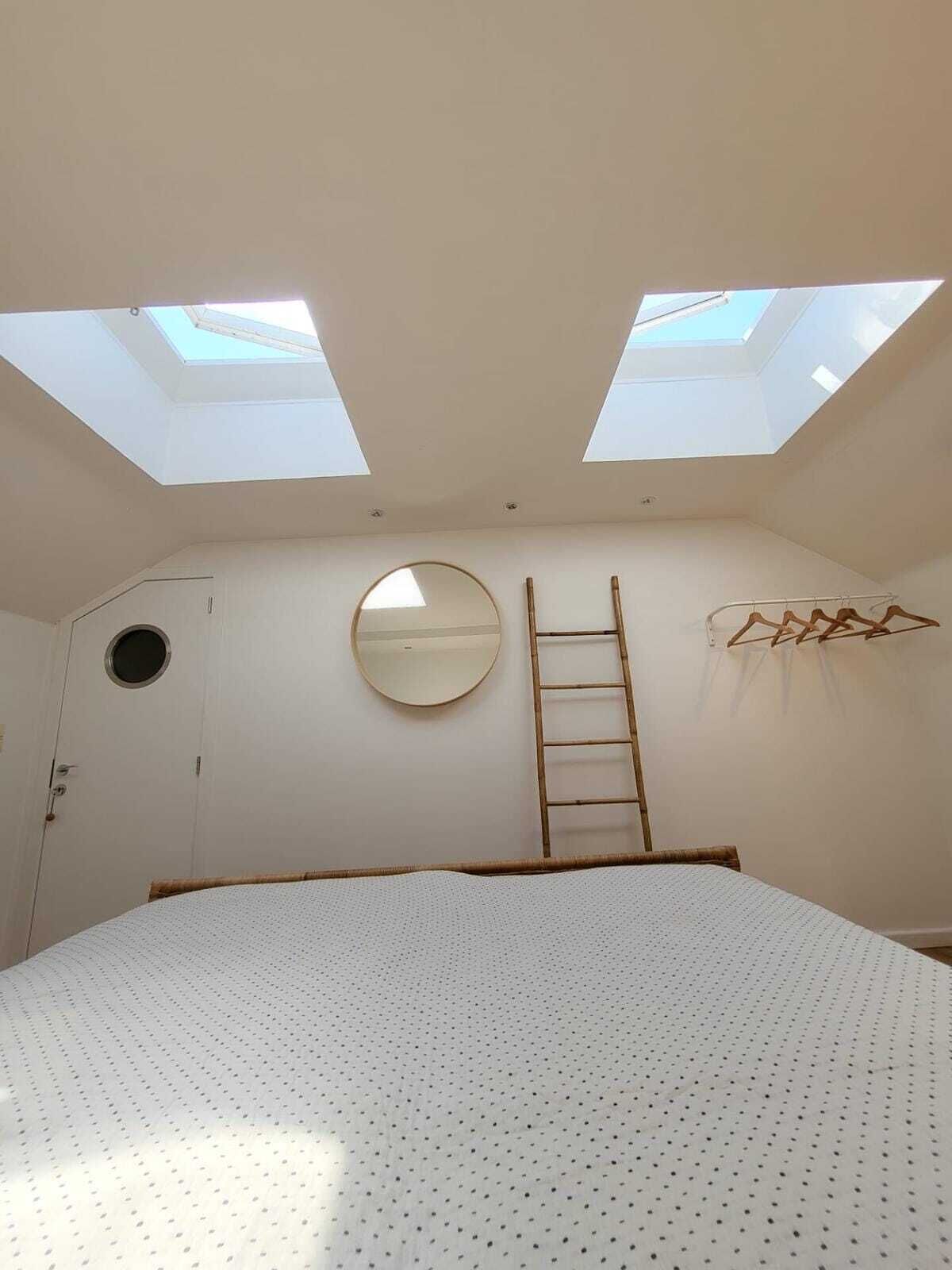 A bedroom with a skylight, a round mirror, a wooden ladder, and a bed with a white, dotted cover.