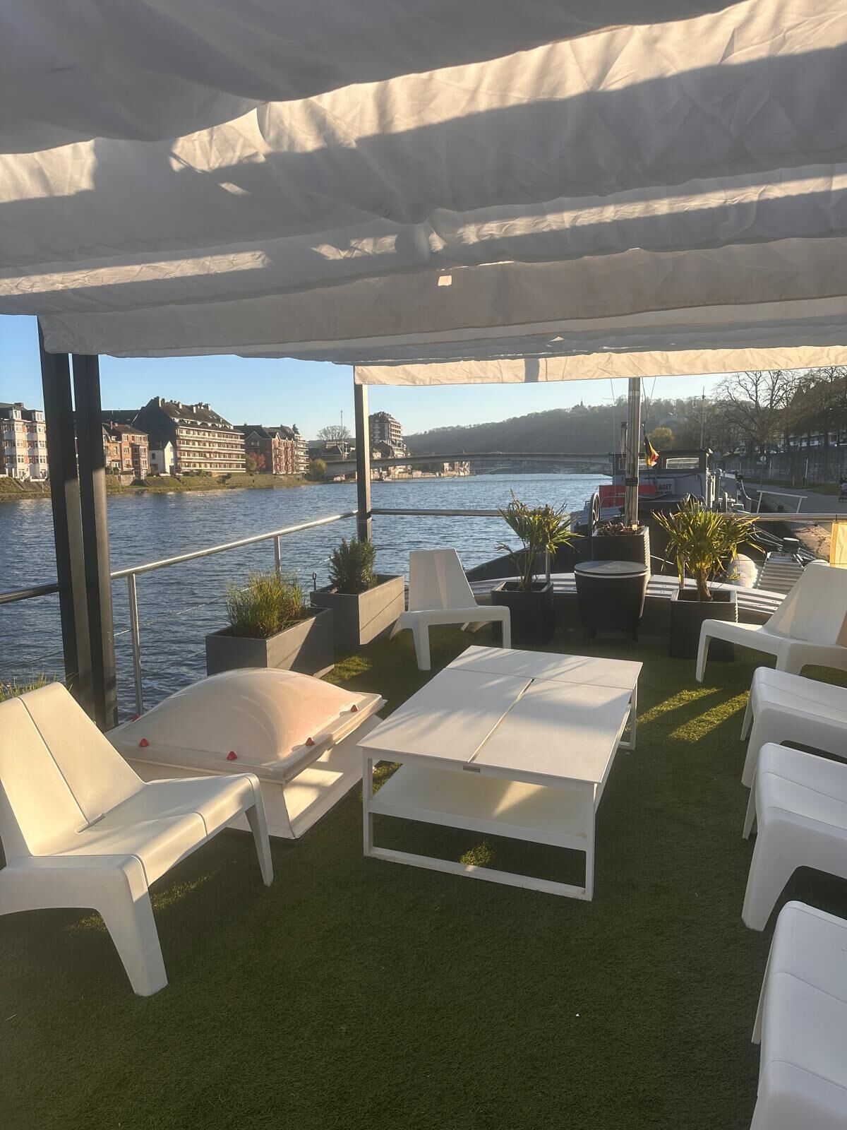 Een terras met witte stoelen en tafels, een gazon en uitzicht op een rivier met gebouwen op de achtergrond.